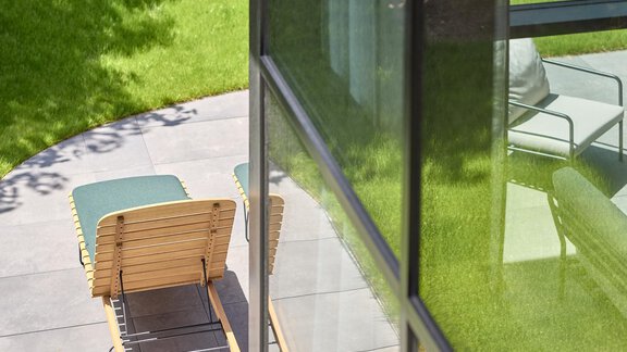 Ein moderner Patiositz mit Blick auf eine grüne Rasenfläche. Die Glaswände des Gebäudes reflektieren das Licht und schaffen eine helle Atmosphäre. - Chalet White Pearl by MYALPS - Sölden
