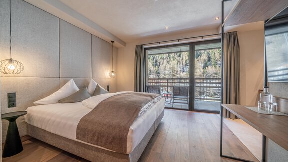 A modern bedroom with a large double bed and wooden floor. Large windows offer a view of the surrounding nature. - Edelweiss echt.charmant. - Sölden