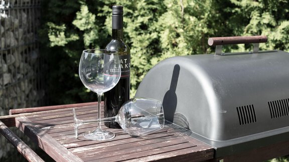 A grill on a wooden table with two wine glasses and a bottle of wine. In the background, green shrubs are visible. - Garni Rustica - Sölden