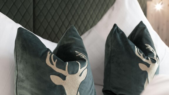 Two stylish cushions in dark green featuring a stag motif. They are placed on a white bed with a textured back wall. - Garni Rustica - Sölden