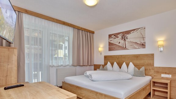 Ein modernes Schlafzimmer mit einem Holzbett und einem Fernseher. Die Wände sind hell, und es gibt große Fenster mit Vorhängen. - Gästehaus Bergkristall - Sölden