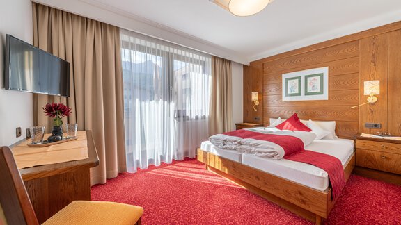 Ein gemütliches Hotelzimmer mit Holzverkleidung und rotem Teppich. Es gibt ein großes Fenster mit Vorhängen und einen TV an der Wand. - Gästehaus Maier - Sölden