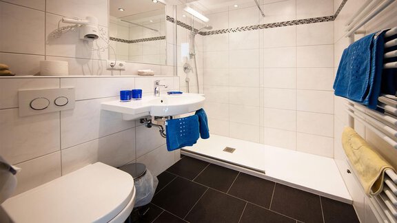 Ein modernes Badezimmer mit einer Dusche und weiß gefliesten Wänden. Es gibt einen Waschbeckenbereich mit blauen Handtüchern und einer eleganten Ausstattung. - Gästehaus Veit Fiegl - Sölden