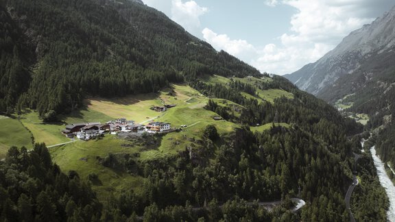 Eine malerische Landschaft mit grünen Hügeln und einem kleinen Dorf. Im Hintergrund sind hohe Berge und ein klarer Himmel zu sehen. - Gasthof Granstein - Sölden
