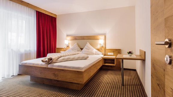 A modern bedroom with a large bed and wooden details. The room has plenty of daylight through a window with curtains. - Haus Madeleine - Sölden