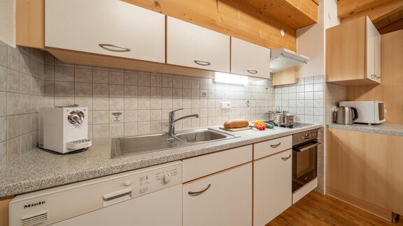 A modern kitchen with wooden shelves and bright cabinets. There is a sink, a stove, and various kitchen appliances. - Haus Nicola - Sölden
