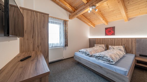 A modern bedroom with wooden beam ceiling and a double bed. The furniture is in a warm wood tone, and there is a television as well as a window looking outside. - Haus Nicola - Sölden