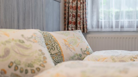 A cozy bedroom with colorful cushions and a wooden panel. In the background, curtains and a window letting in daylight can be seen. - Haus Nicola - Sölden