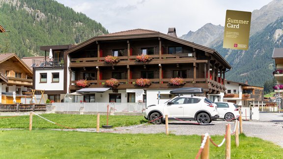Ein schönes Ferienhaus in den Alpen mit vielen Blumen auf dem Balkon. Im Vordergrund steht ein Auto auf einem Parkplatz. - Hotel Garni Maria Theresia - Sölden