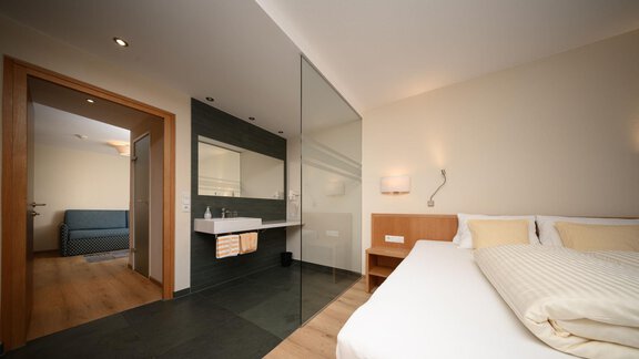 Ein modernes Zimmer mit einem Doppelbett und einem Badezimmer mit Glaswand. Im Hintergrund ist ein weiteres Zimmer mit einem Sofa zu sehen. - Hotel Garni Maria Theresia - Sölden