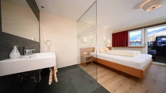 Ein modernes Schlafzimmer mit einem großen Fenster und Aussicht auf die Berge. Das Bad ist durch eine Glaswand vom Raum getrennt. - Hotel Garni Maria Theresia - Sölden