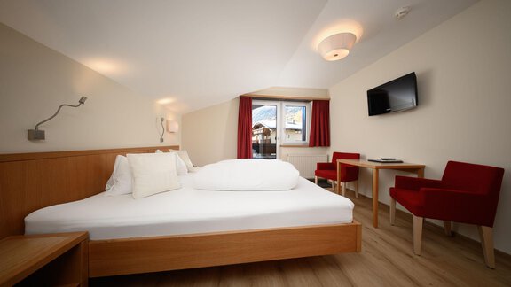 Ein modernes Schlafzimmer mit einem Doppelbett und einem gemütlichen Tisch. Rote Stühle und ein Flachbildfernseher ergänzen die Einrichtung. - Hotel Garni Maria Theresia - Sölden