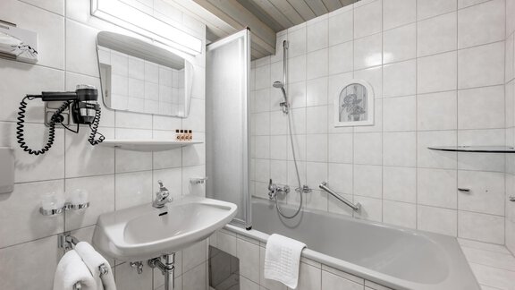 Ein modernes Badezimmer mit weißen Fliesen, einer Badewanne und einer Dusche. Es gibt ein Waschbecken mit Spiegel und Handtüchern. - Hotel Sölderhof - Sölden