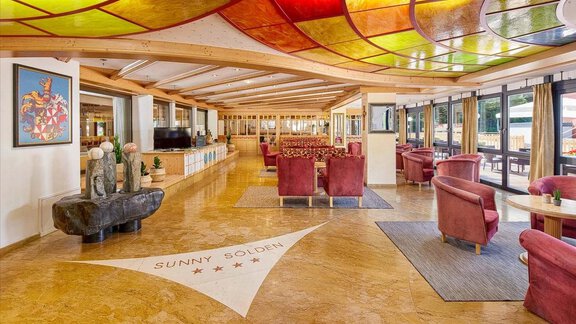 An elegant interior with red sofas and large windows. An artistic glass roof lets warm light in. - Hotel Sunny Sölden - Sölden