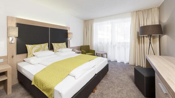 A modern hotel room with two beds and a variety of cushions in yellow and green. The room features a window with curtains and a cozy seating area. - Hotel Tyrolerhof - Sölden
