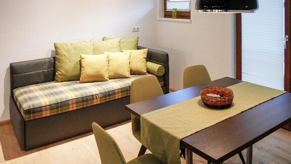 A cozy living room with a gray sofa and green cushions. A wooden table with four chairs stands beside it, decorated with a fruit bowl. - Lutonia Appartements Sölden - Sölden