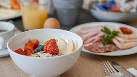 A bowl of fresh fruit, yogurt, and oatmeal is placed on a table. In the background, other breakfast dishes such as sausage and a drink can be seen. - Pension Charly - Sölden