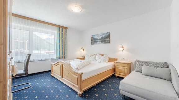 A cozy bedroom with a wooden bed and a sofa bed. Large windows provide plenty of daylight and offer a view of the surroundings. - Pension Charly - Sölden