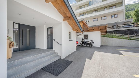 A modern building with an entrance area and a staircase. In the background, additional apartment blocks and a motorcycle cover are visible. - Pension Charly - Sölden