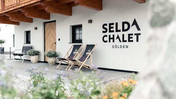 Ein modernes Chalet mit dem Schriftzug "SELDA CHALET" in Sölden. Es gibt bequeme Lounge-Stühle und Blumen vor der Eingangstür. - Selda Chalet - Sölden