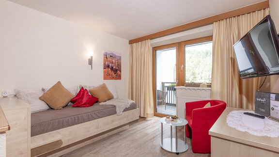 A cozy living room with a single bed - which can also be used as a sofa. The large windows offer a view outside and the room is bright and comfortably designed. - Trofana Appartements - Sölden
