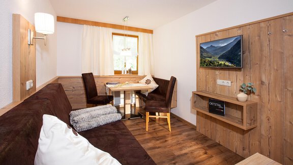 A cozy living space with wood paneling and a modern television. A table with chairs and a comfortable single bed - which can also be used as a sitting couch - invite you to relax. - Trofana Appartements - Sölden