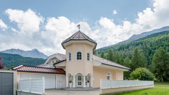 Ein modernes Haus mit einem runden Turm und einem roten Satteldach. Im Hintergrund sind grüne Berge und ein blauer Himmel zu sehen. - Villa Castelbell - Sölden