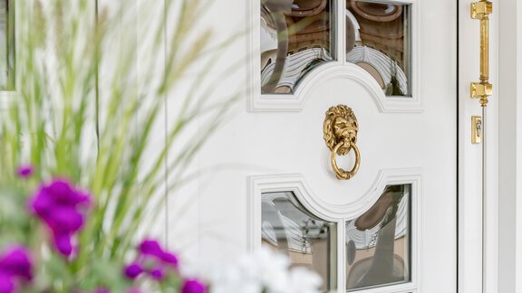 Eine elegante weiße Tür mit einem goldenen Türklopfer in Form eines Löwen. Um die Tür herum blühen bunte Blumen. - Villa Castelbell - Sölden