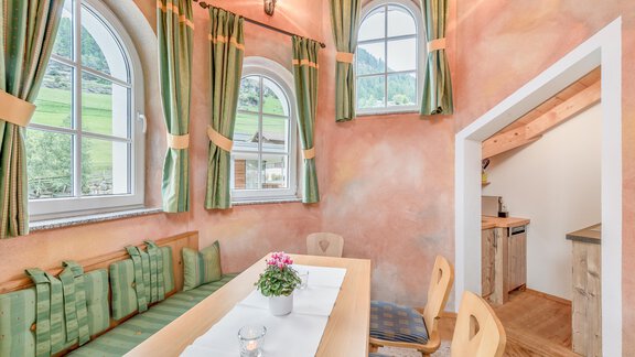 Ein helles, einladendes Zimmer mit einem langen Tisch und gemütlichen Sitzgelegenheiten. Die Wände sind in sanften Farben gehalten, und es gibt große Fenster mit grünen Vorhängen. - Villa Castelbell - Sölden