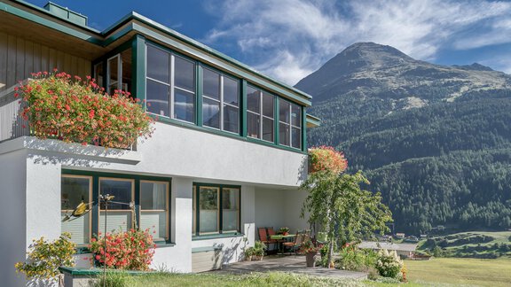 Ein modernes Haus mit großen Fenstern und blühenden Pflanzen. Im Hintergrund erstrecken sich grüne Berge unter einem blauen Himmel. - zuckerhütl NATURAPPART - Sölden