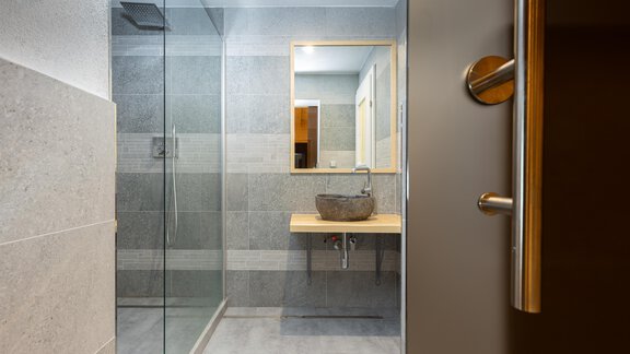 A modern bathroom with a glass shower and a bright wood vanity. The walls are in neutral gray tones, and a large mirror provides light. - Garni Stefani - Vent