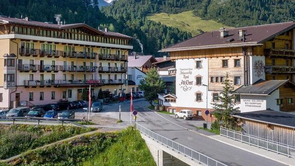 Zwei Hotels in einer malerischen Berglandschaft. Grüne Hügel und eine ruhige Straße umgeben die Gebäude. - Hotel Vent - Vent