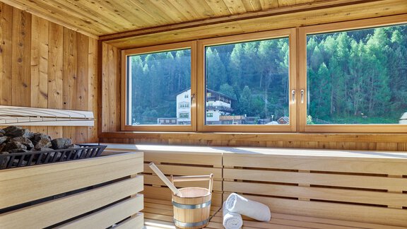 Eine moderne Sauna mit Holzwänden und großen Fenstern, die eine schöne Aussicht auf den Wald bietet. Im Vordergrund sind eine Saunabank, Steine und ein Holzbehälter zu sehen. - Hotel Vent - Vent