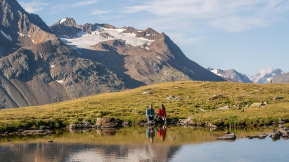Eine malerische Berglandschaft mit schneebedeckten Gipfeln und einem ruhigen See. Zwei Personen sitzen am Ufer und genießen die Aussicht. - Hotel Vent - Vent