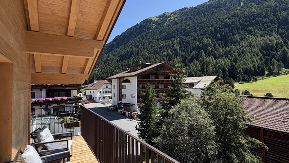 A balcony with comfortable seating offers a view of green mountains and picturesque houses. The sky is clear and blue, creating a warm atmosphere. - Mein Weisskugel - Vent