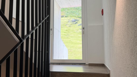 A staircase with wooden flooring leads to a glass door. Light streams through the window and the railing is made of black metal. - Mein Weisskugel - Vent