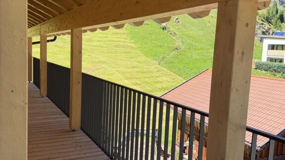 A balcony with wooden paneling and a view of a green meadow. In the background, there are distant buildings and a clear view of the sky. - Mein Weisskugel - Vent