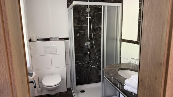 A modern bathroom with a glass shower and dark tiles. There is a sink with towels and a toilet. - Mein Weisskugel - Vent