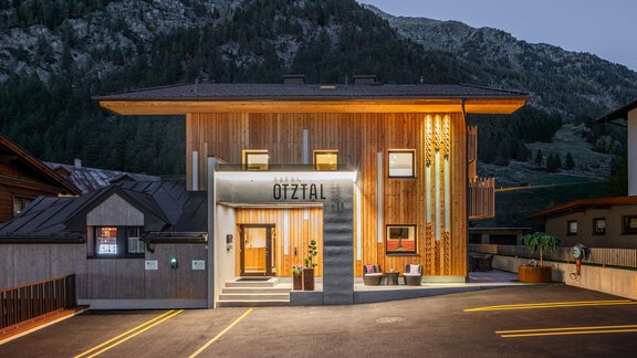 A modern building with a wooden facade in a picturesque mountain village. The facade is pleasantly illuminated and there are parking spaces in the foreground. - Garni Ötztal ROOMS & APARTS - Zwieselstein