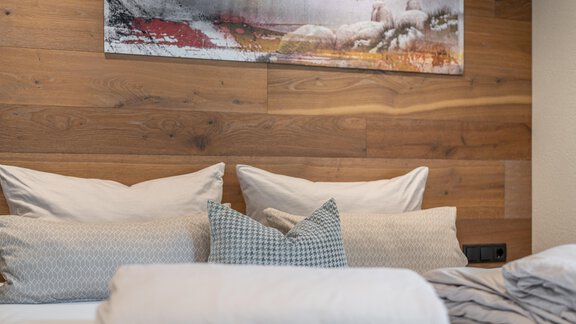 A cozy bedroom with a wooden wall design and several pillows on the bed. Above the bed hangs a colorful piece of artwork. - Garni Ötztal ROOMS & APARTS - Zwieselstein