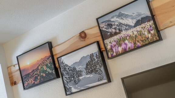 Three framed pictures hang on a wooden strip on the wall. The photos depict a landscape with a sunset, snow-covered mountains, and blooming flowers. - Garni Ötztal ROOMS & APARTS - Zwieselstein