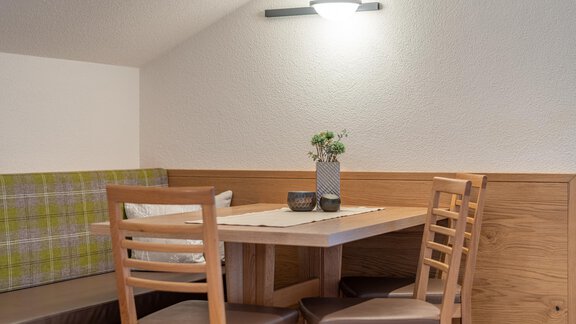 A cozy dining area with a wooden chair and a table. On the table is a vase with flowers and there is a bench in the background. - Garni Ötztal ROOMS & APARTS - Zwieselstein