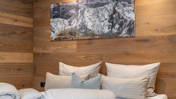 A cozy bedroom with wooden panels and white cushions. On the wall hangs a picture of snow-covered mountains. - Garni Ötztal ROOMS & APARTS - Zwieselstein