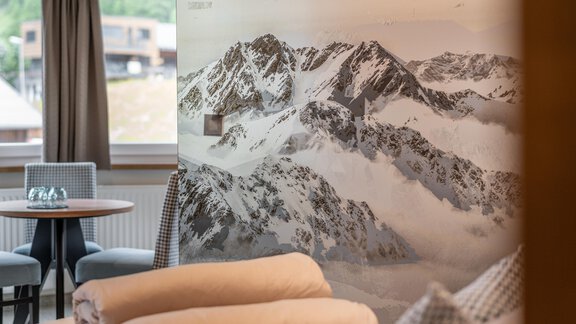 A modern room with wall design that features majestic mountains. In the foreground, cozy cushions and a table with chairs are visible. - Garni Ötztal ROOMS & APARTS - Zwieselstein