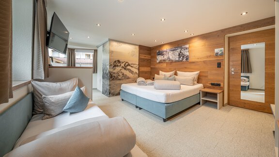 A modern hotel room with a comfortable bed and a sofa. The walls are decorated with wooden paneling, and there is a television as well as a window with a view. - Garni Ötztal ROOMS & APARTS - Zwieselstein