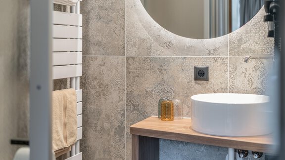 Ein modernes Badezimmer mit eleganten Fliesen und einem runden Spiegel. Darunter befindet sich ein Waschbecken aus weißem Keramik und ein Holzregal. - Garni Ötztal ROOMS & APARTS - Zwieselstein