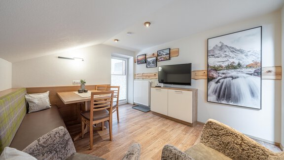 A cozy living space with a dining table, a television, and wall decoration. Large windows provide plenty of light and an inviting atmosphere. - Garni Ötztal ROOMS & APARTS - Zwieselstein