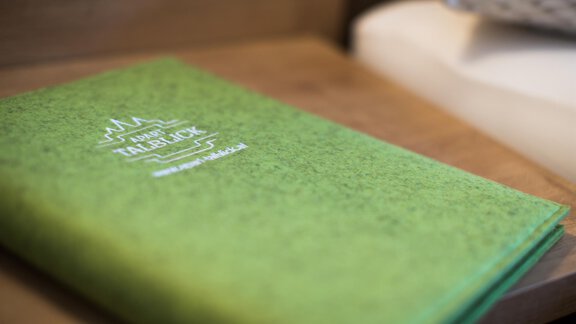 Ein grünes Notizbuch liegt auf einem Holztisch. Es handelt sich um eine Einladung oder ein Programm mit einem klaren Logo. - Apart Talblick - Sölden