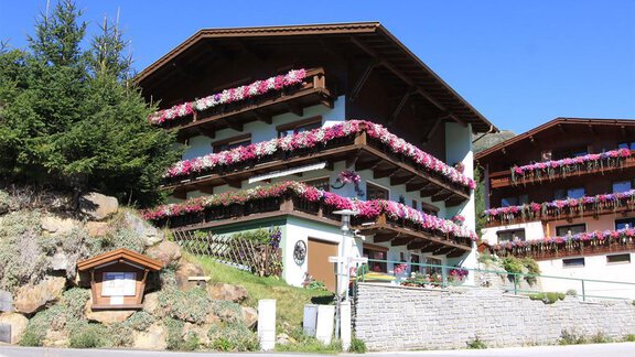 Ein traditionelles Alpenhaus mit bunten Blumen auf den Balkonen. Umgeben von grünen Bäumen und einem klaren blauen Himmel. - Gästehaus Veit Fiegl - Sölden