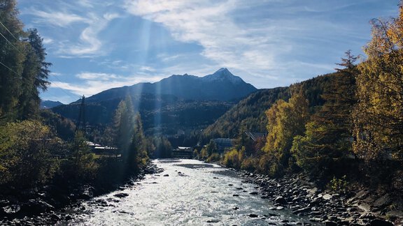 Ein klarer Fluss fließt durch eine herbstliche Landschaft mit bunten Bäumen. Im Hintergrund sind Berge unter einem strahlend blauen Himmel zu sehen. - Granbichlhof - Sölden